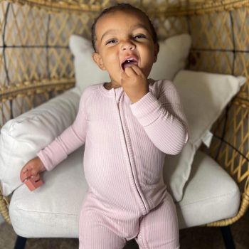 A tiny baby sits in a wicker chair with her mouth open.